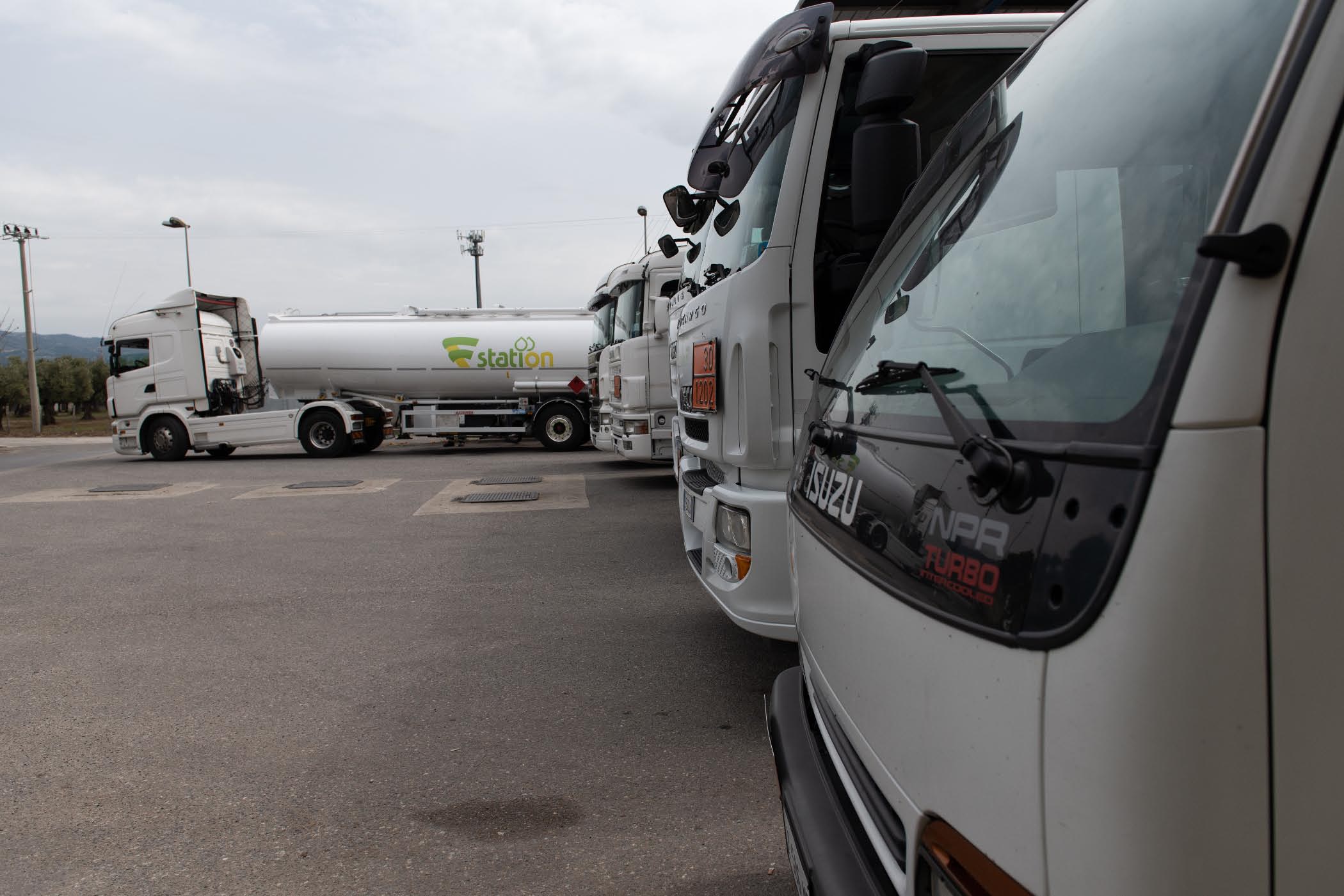 Autocisterna bianca con logo station e camion cisterna in fila con pannelli arancioni ADR, tra cui un Isuzu NPR Turbo